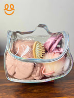 Clear toiletry bag with pink towel, wooden hairbrush, bath sponge, and pink fabric items on wooden surface