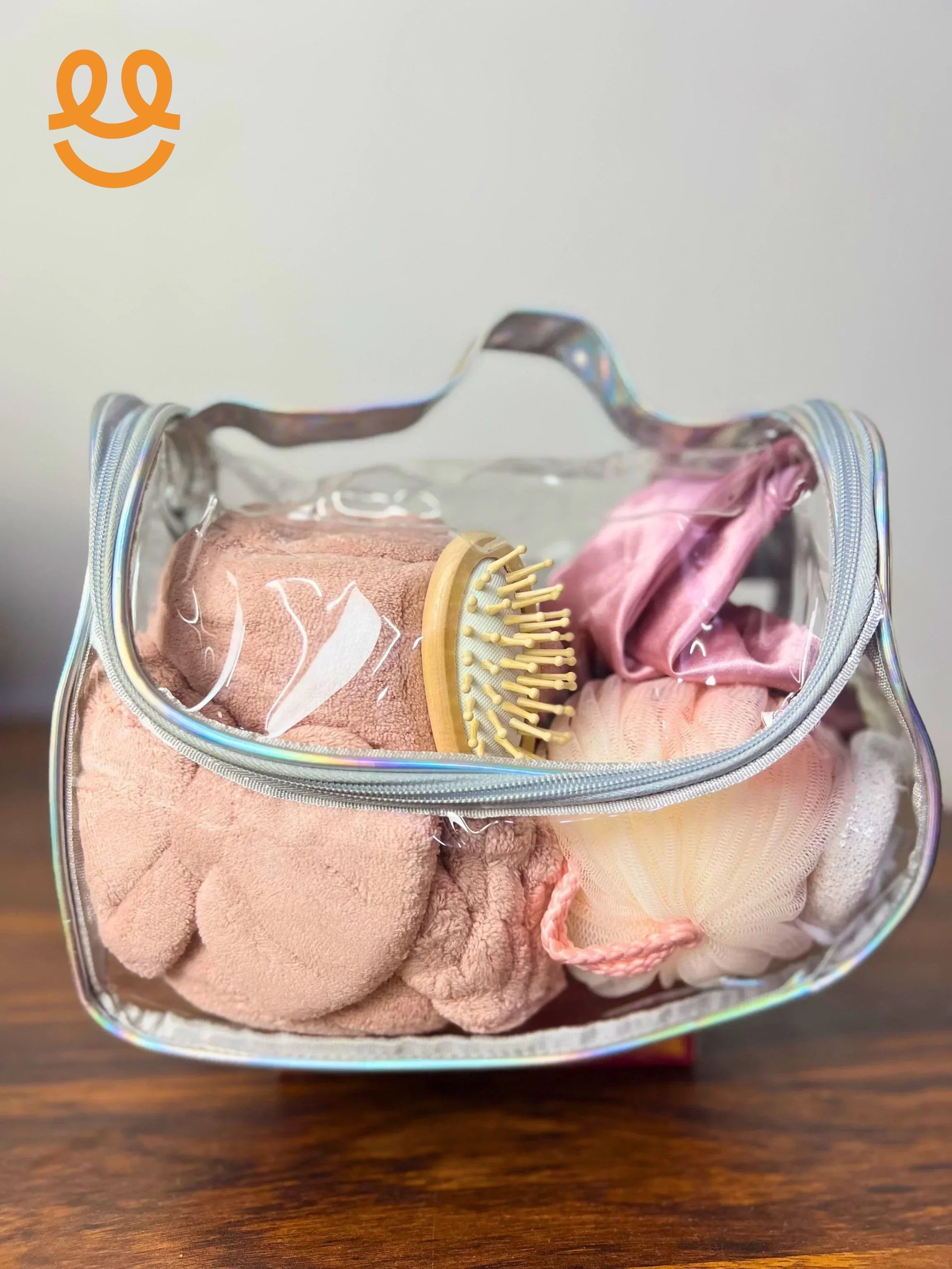 Clear toiletry bag with pink towel, wooden hairbrush, bath sponge, and pink fabric items on wooden surface