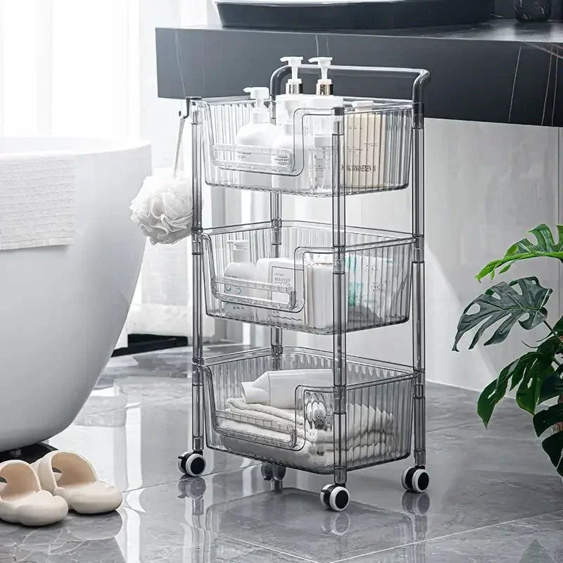 Three-tier clear plastic rolling bathroom storage cart with bottles, towels, and toiletries on gray tiled floor