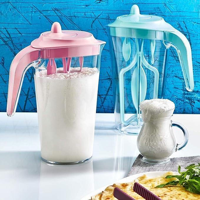 Two transparent milk frothers with pink and blue lids, one filled with frothy milk, next to a glass jug of frothed milk on white surface