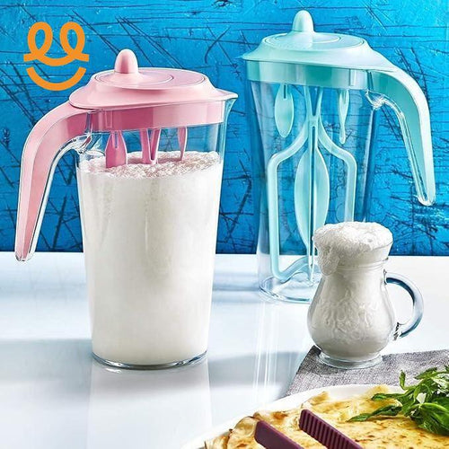 Two transparent milk frothers with pink and blue lids, one filled with frothy milk, next to a glass jug of frothed milk on white surface