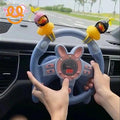 Hands holding a blue toy steering wheel with pink bunny ears and two yellow duck decorations inside a car dashboard