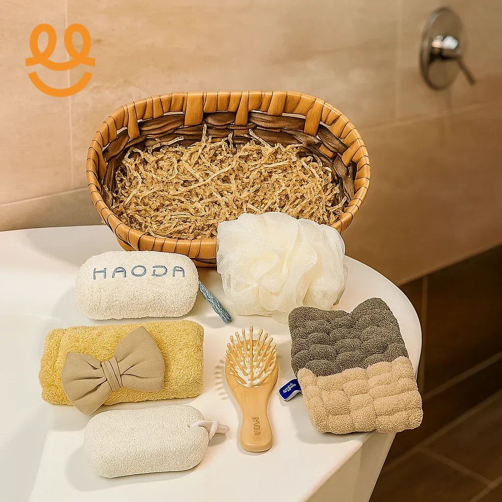Wicker basket with shredded filler and bathroom essentials including sponge, bath pouf, wooden hairbrush, towel with bow, and exfoliating gloves on white surface