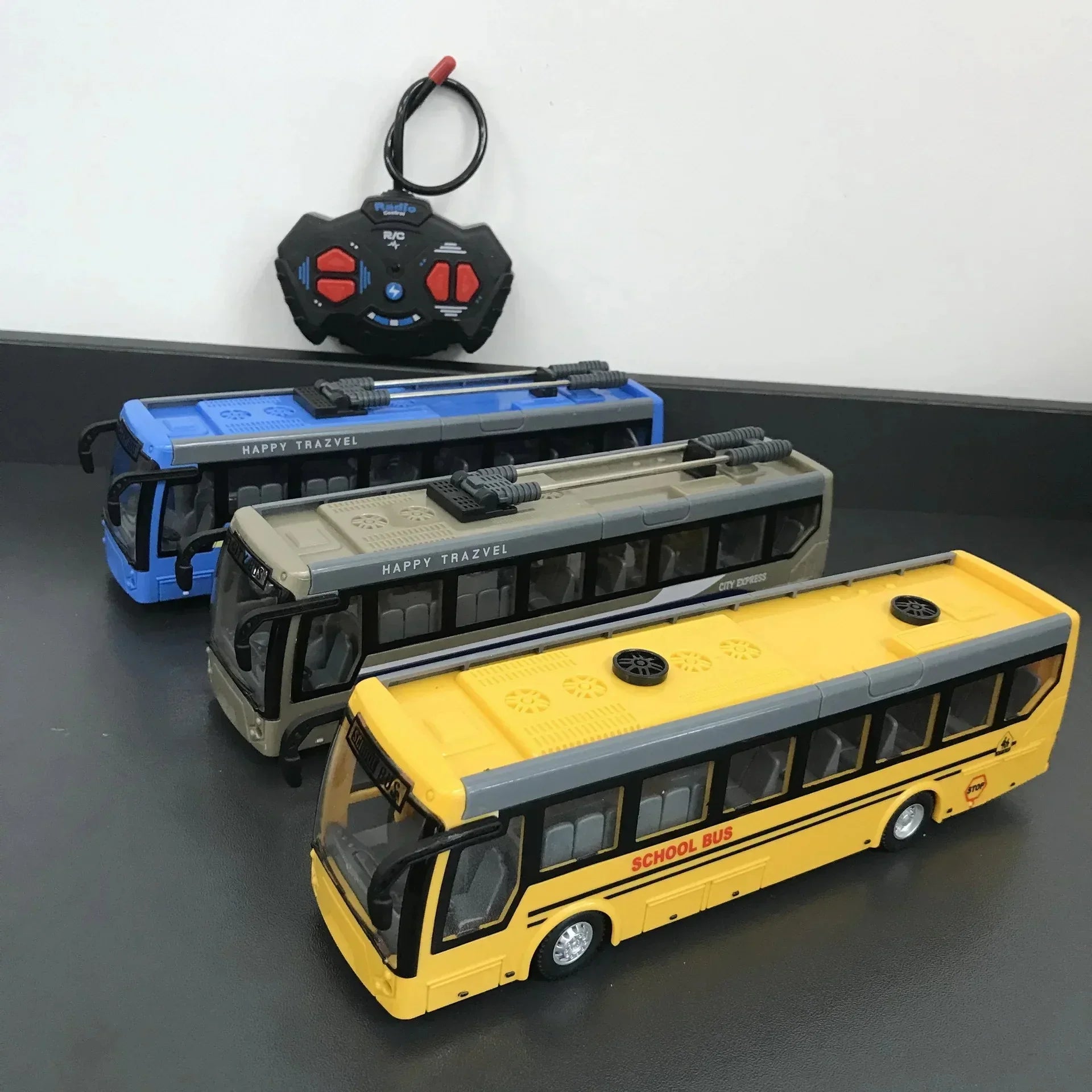 Three remote control toy buses in yellow, beige, and blue with a black and red remote on a gray surface
