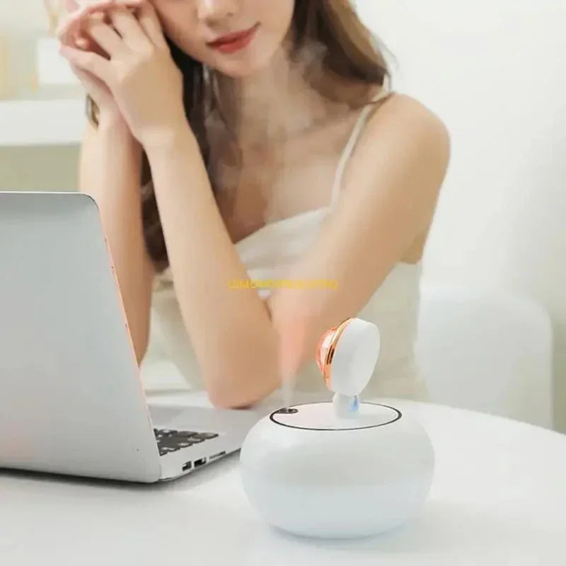 Woman using a white facial mist humidifier on a desk next to a laptop in a bright room