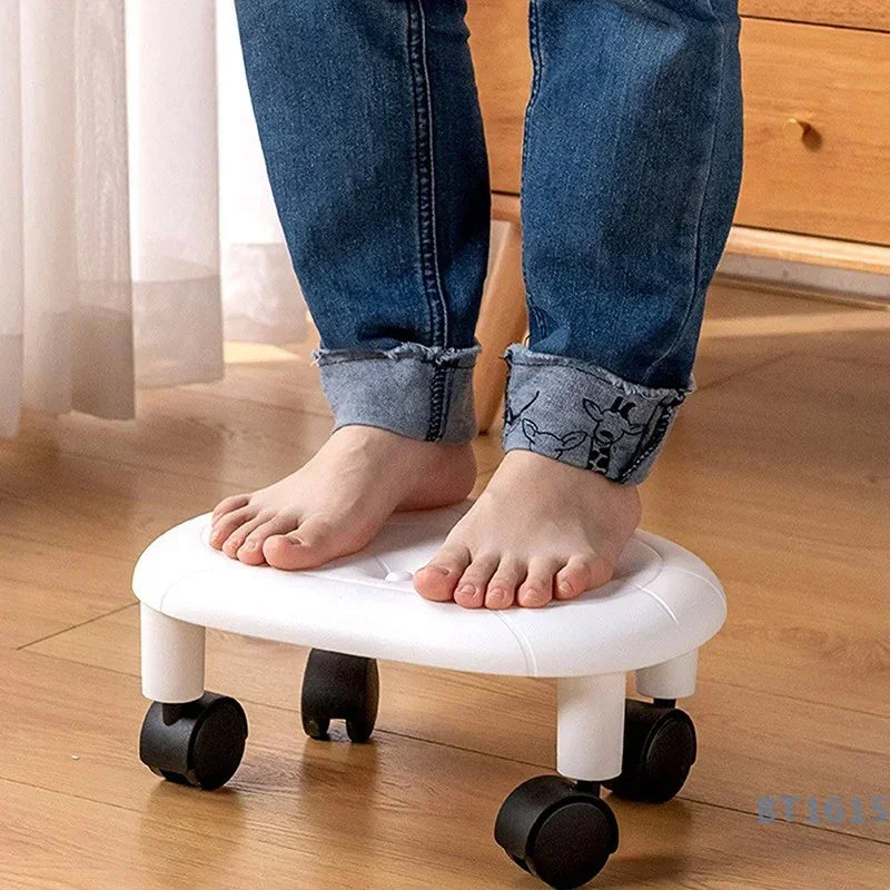 Bare feet standing on small white rolling footrest with four black wheels on wooden floor