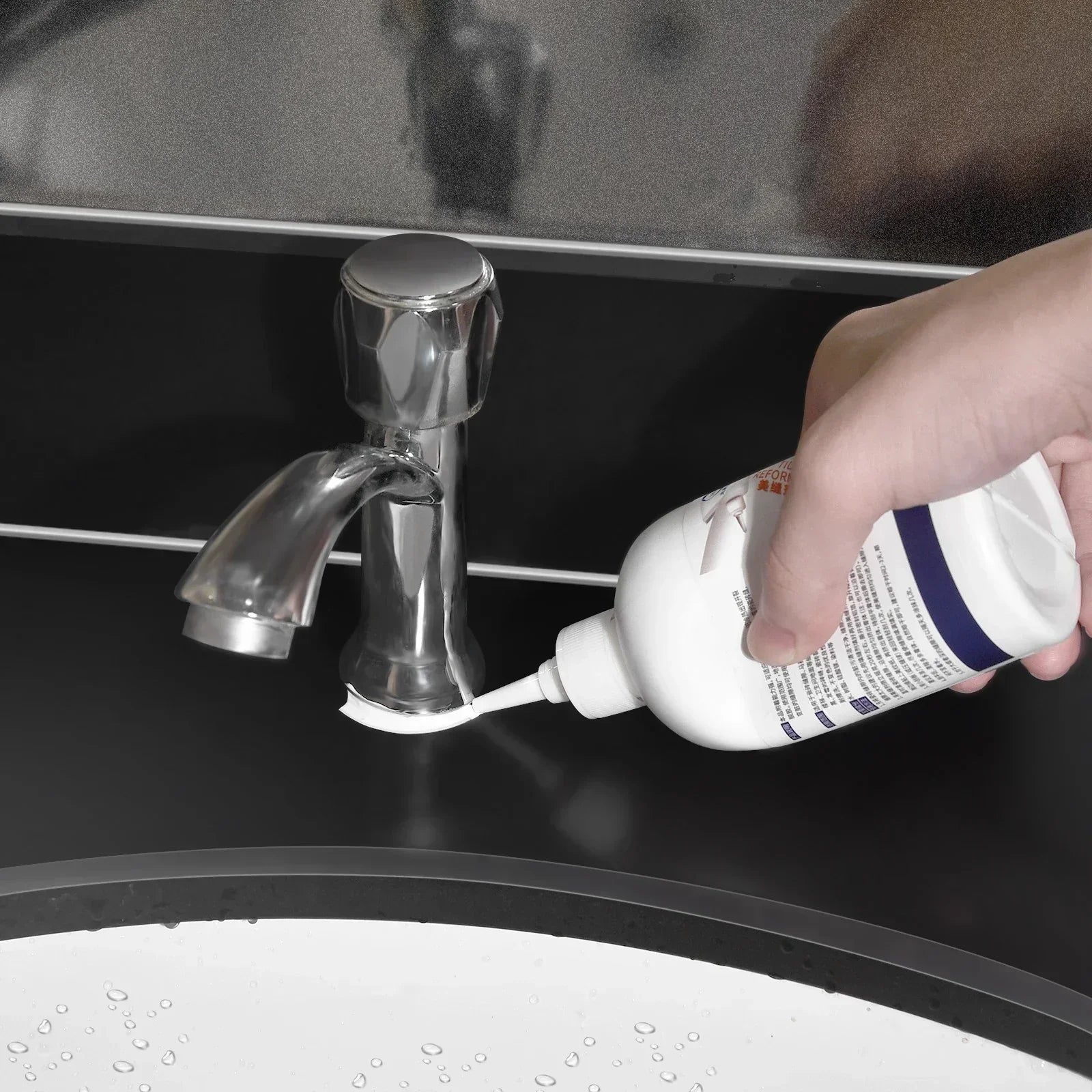 Hand applying white sealant around base of silver faucet on black bathroom sink countertop