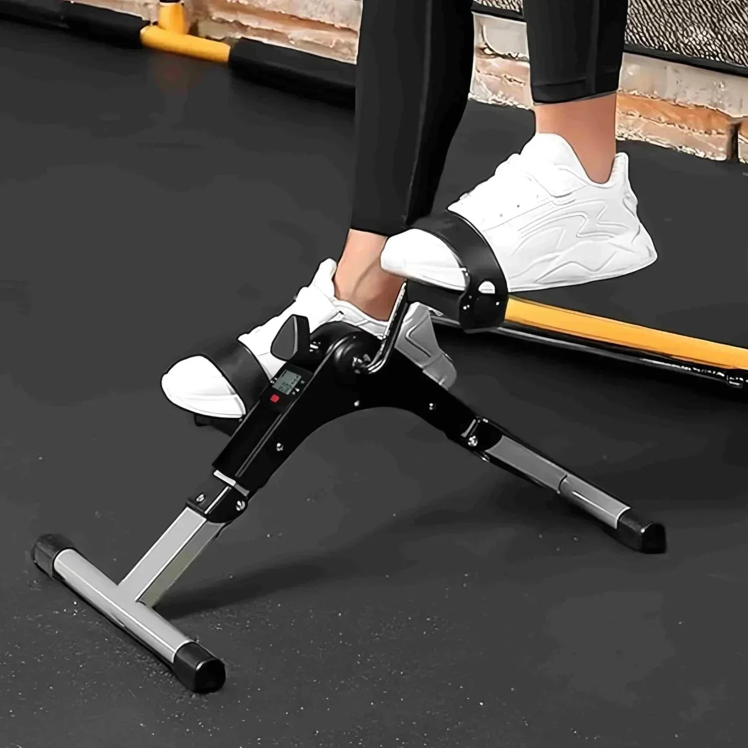 Person using a compact mini exercise bike pedal exerciser with white sneakers on black gym floor