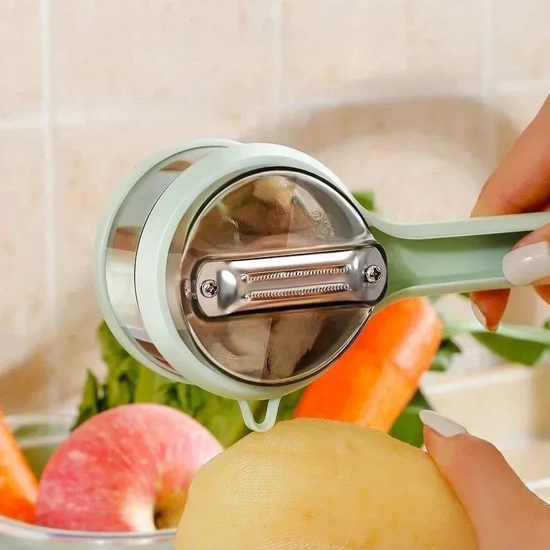 Hand using a handheld rotary vegetable peeler to peel a potato with carrots and an apple in the background