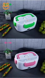 Two electric lunch boxes with handles, one green and one pink, on a dark surface with grapes, sausages, and a small plant