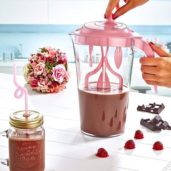 Clear pitcher with pink lid and handle filled with chocolate milkshake, next to mason jar with pink straw, chocolate pieces, and strawberries on white table