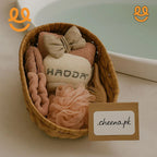 Wicker basket with beige bath accessories including headband, bow, loofah, wooden brush, and HADDA branded towel