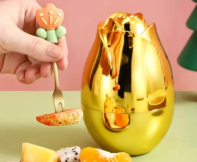 Hand holding a floral-handled fork with fruit slice near a shiny gold rose-shaped container on a green and pink background