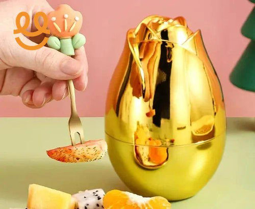 Hand holding a floral-handled fork with fruit slice near a shiny gold rose-shaped container on a green and pink background