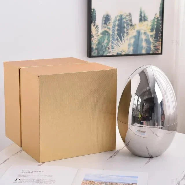 Chrome metallic egg-shaped decorative object beside plain brown cardboard box on marble table with cactus framed art