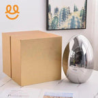Chrome metallic egg-shaped decorative object beside plain brown cardboard box on marble table with cactus framed art