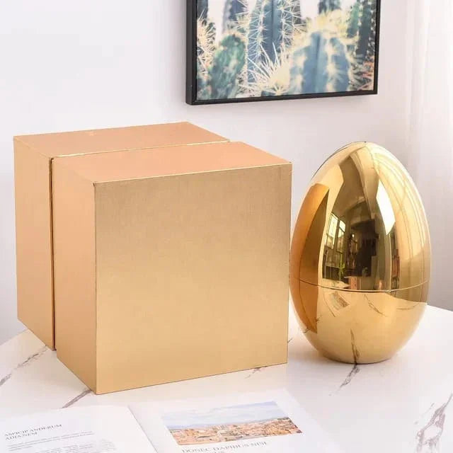 Gold decorative egg and two gold gift boxes on white marble table under cactus wall art