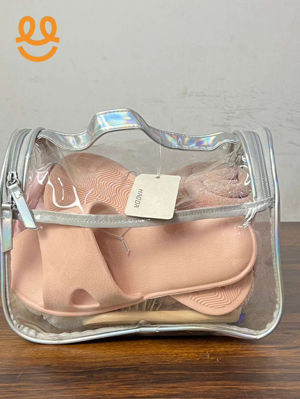 Clear iridescent travel bag containing pink slip-on sandals and a matching pink towel