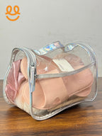 Clear travel bag with iridescent trim holding pink slide sandals and soft pink fabric on wooden surface