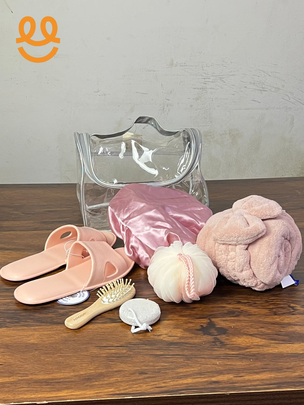 Clear toiletry bag with pink slippers, satin shower cap, bath sponge, wooden hairbrush, exfoliating pad, and pink towel on wood