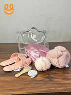 Clear toiletry bag with pink slippers, satin shower cap, bath sponge, wooden hairbrush, exfoliating pad, and pink towel on wood