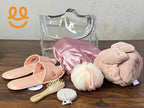 Clear toiletry bag with pink slippers, satin shower cap, bath sponge, wooden hairbrush, pumice stone, and rolled pink towel