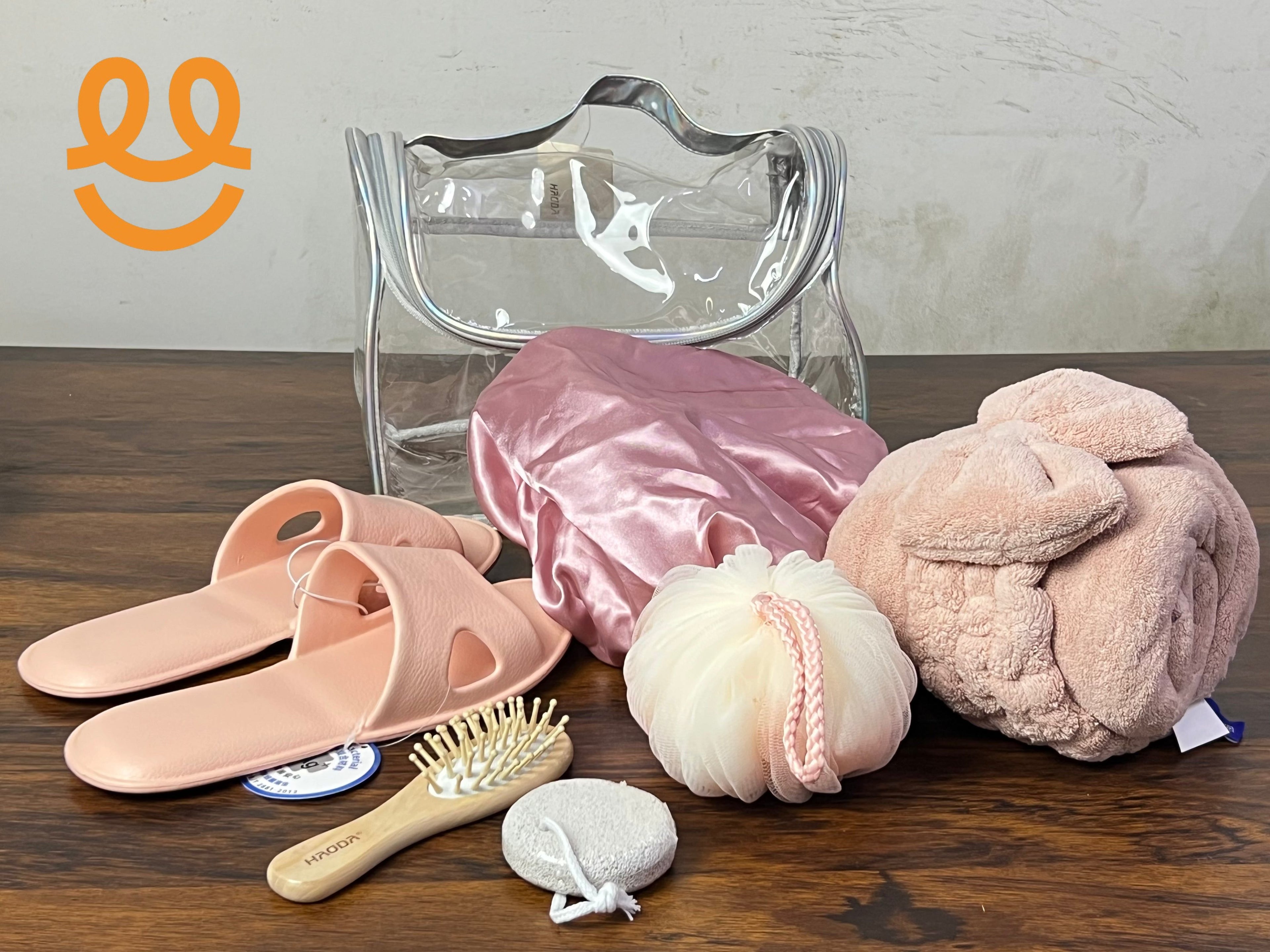 Clear toiletry bag with pink slippers, satin shower cap, bath sponge, wooden hairbrush, pumice stone, and rolled pink towel