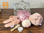 Clear toiletry bag with pink slippers, satin shower cap, pink towel, hairbrush, bath sponge and pumice stone on wood