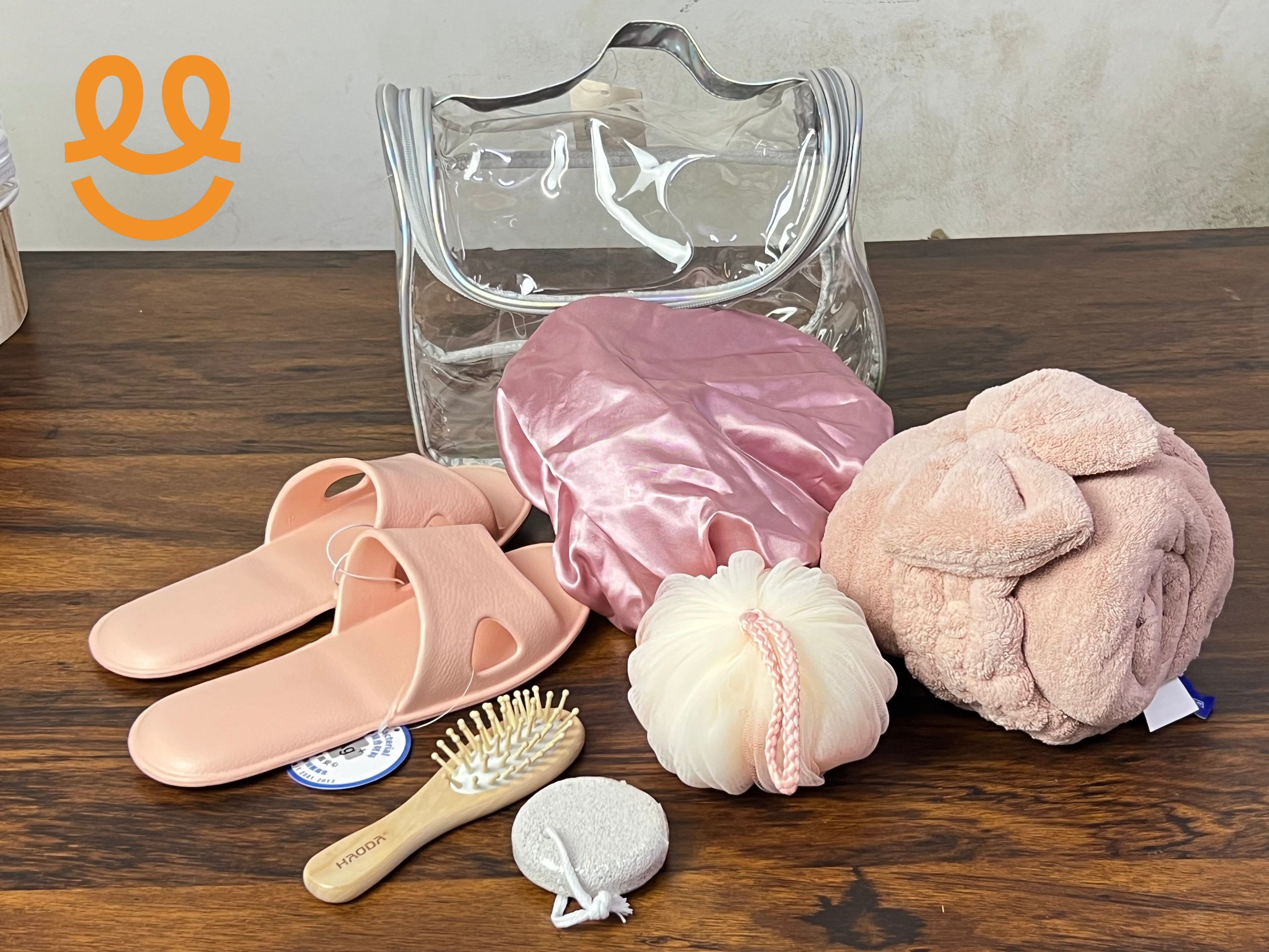 Clear toiletry bag with pink slippers, satin shower cap, pink towel, hairbrush, bath sponge and pumice stone on wood