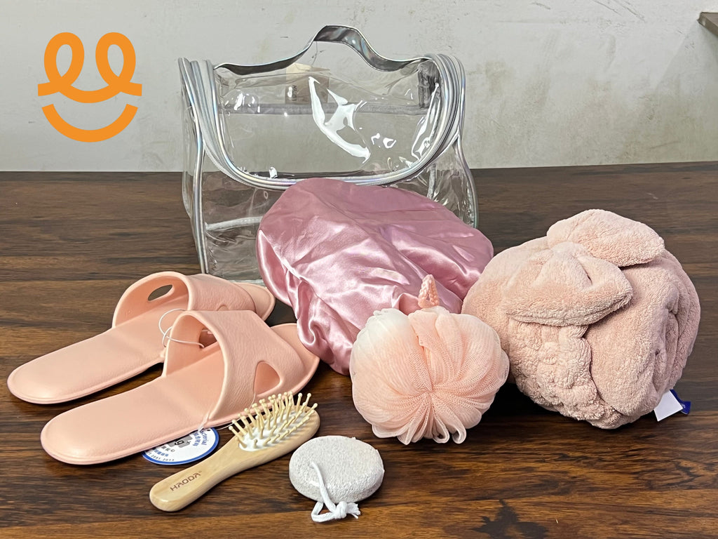 Clear cosmetic bag with pink slippers, satin shower cap, bath sponge, hairbrush, pumice stone, and plush towel on wooden surface