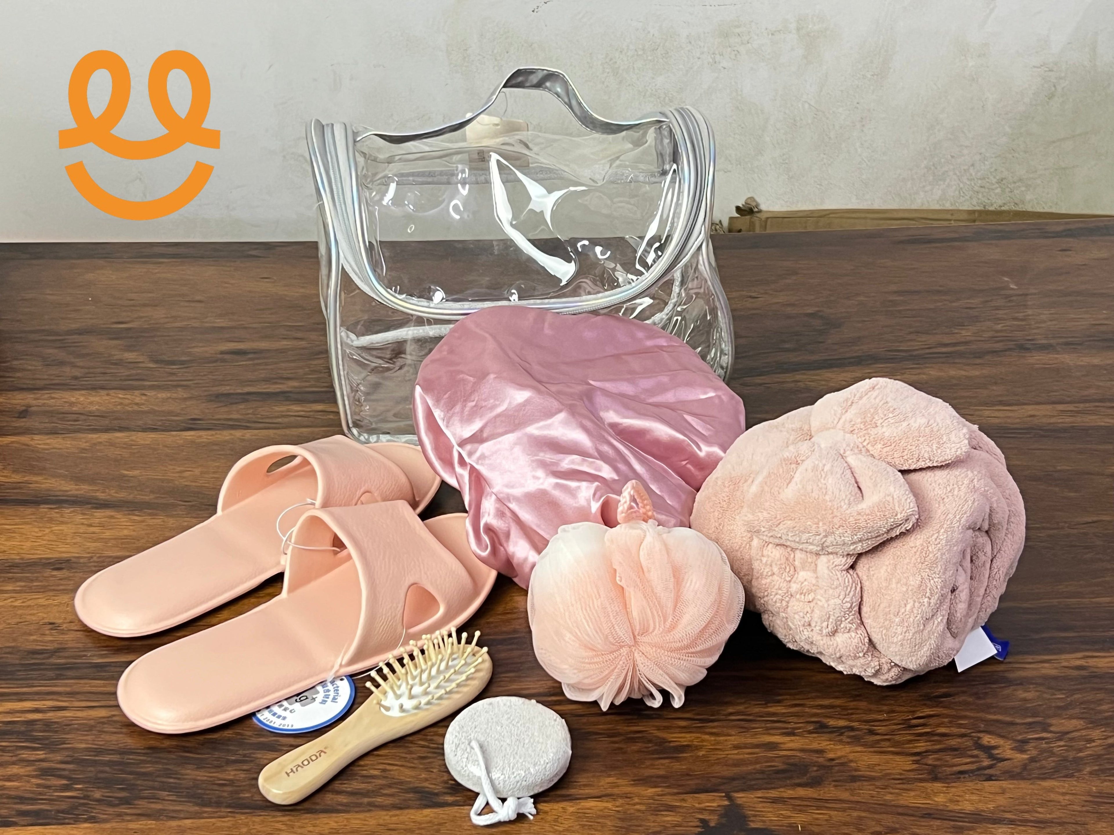 Clear toiletry bag with pink spa slippers, satin bonnet, bath sponge, hairbrush, pumice stone, and soft towel on wooden surface