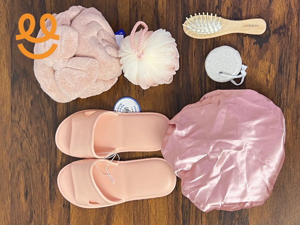 Pink bath slippers, shower cap, bath pouf, headband, hairbrush, and pumice stone on wooden floor