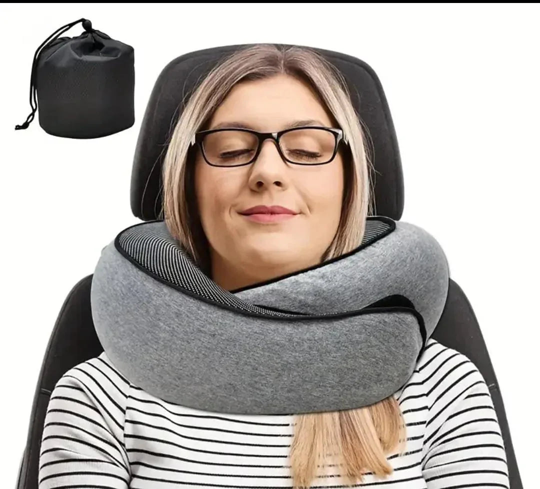 Woman with glasses wearing a gray memory foam travel neck pillow, sitting on black chair, black storage bag