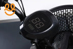 Close-up of electric bike handlebar with digital display showing speed and battery level, black basket in background