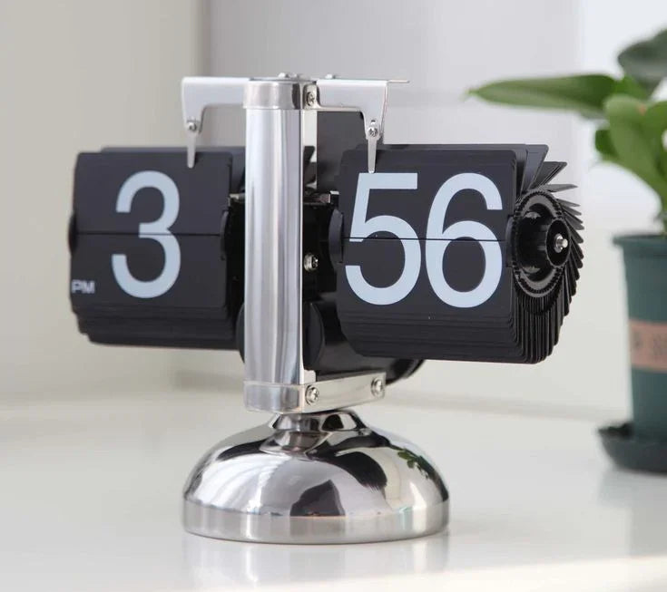 Retro black flip clock showing 3:56 PM with metallic base on white surface and plant in background