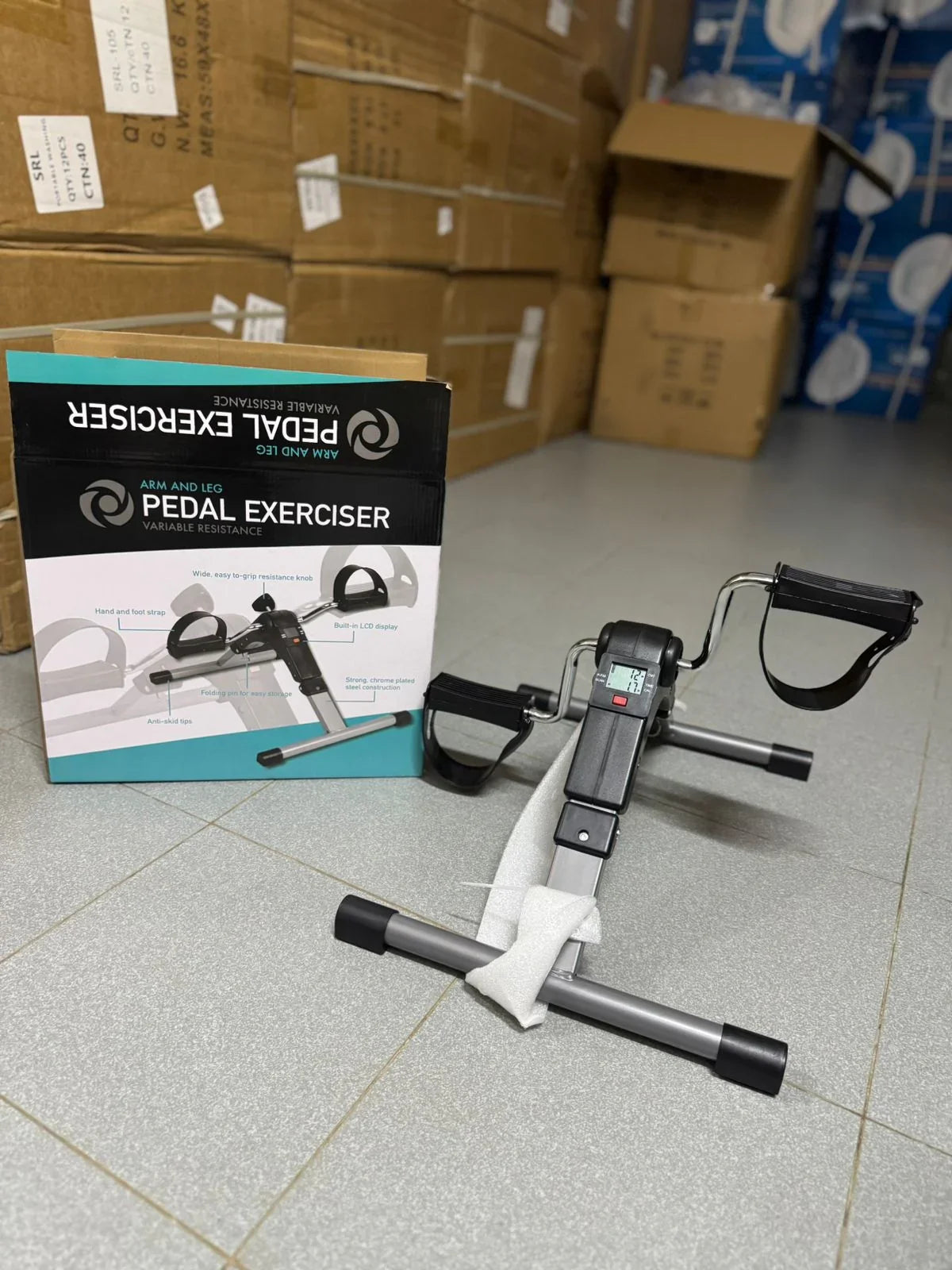 Arm and leg pedal exerciser on gray floor next to its box and stacked cardboard packages