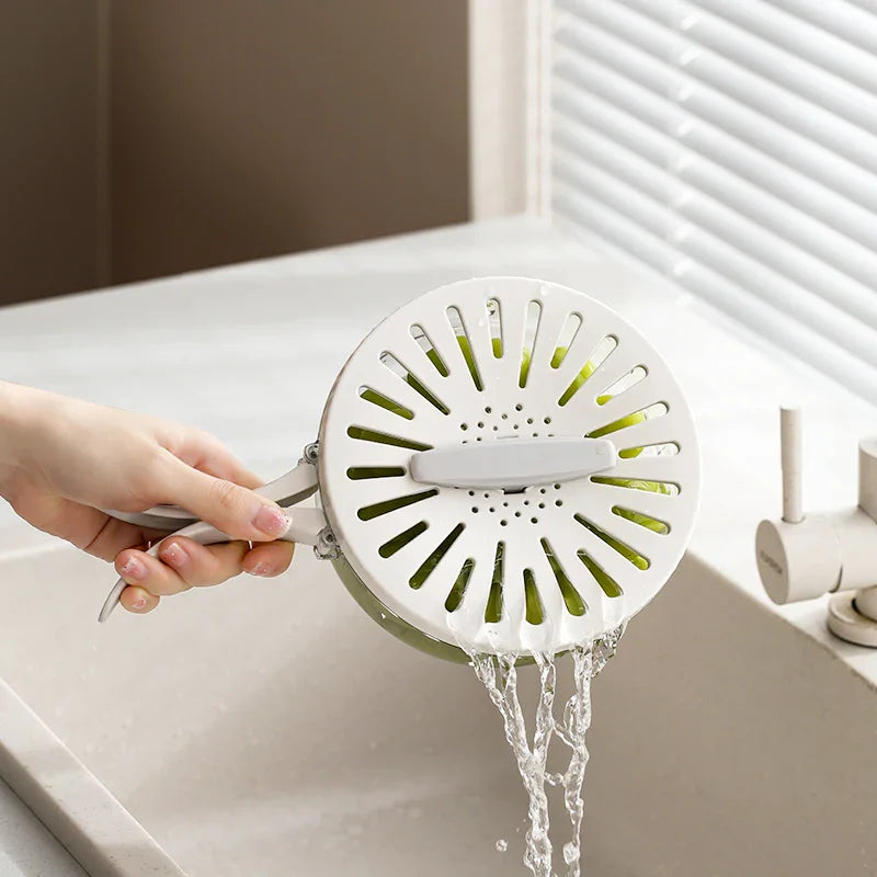 Hand holding white vegetable strainer draining water over kitchen sink with faucet and window blinds