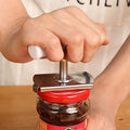 Hand using a jar opener tool to twist open a red-lidded glass jar on wooden surface
