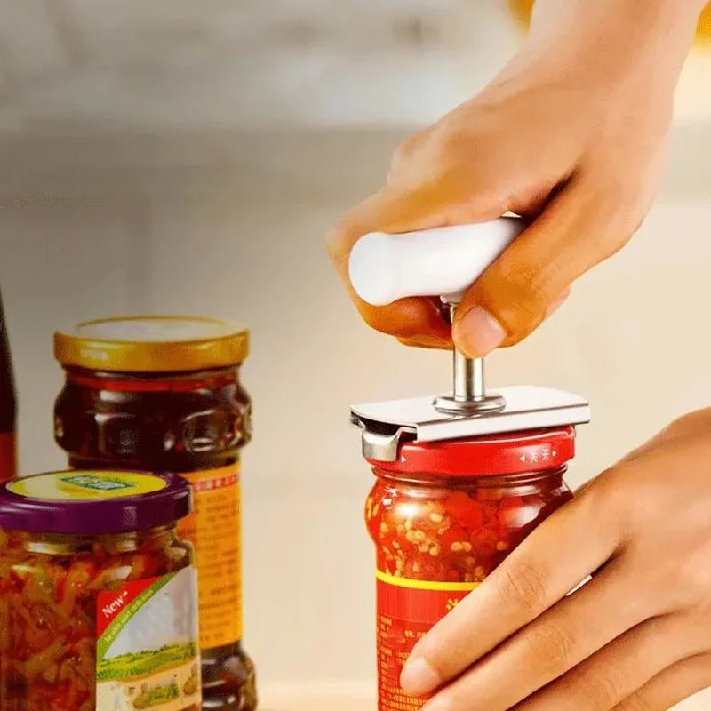 Hand using a white kitchen tool to open a jar of red chili sauce on a countertop with other jars nearby