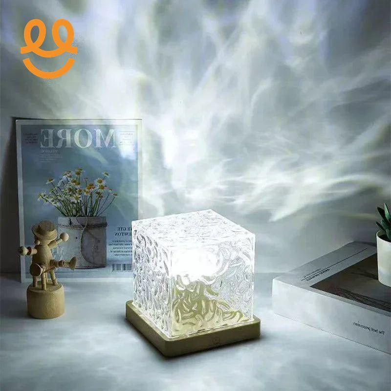 Decorative glass cube lamp emitting soft light with artistic light patterns on a tabletop with a wooden figure and framed flower poster