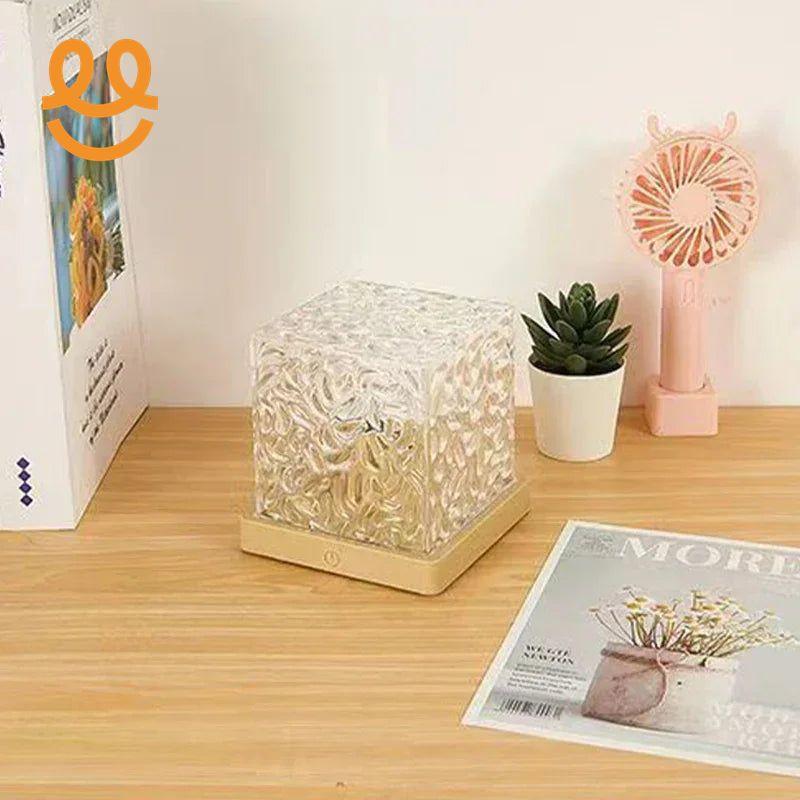 Decorative textured glass cube light on wooden surface with potted succulent, pink fan, and magazine